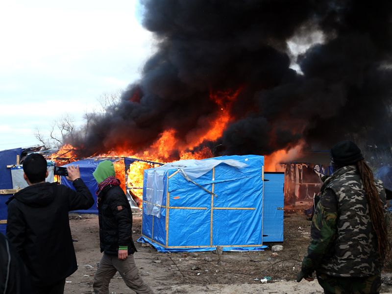 Migrants look on as a hut burns.