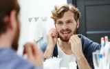 A man flosses his teeth in the bathroom mirror.
