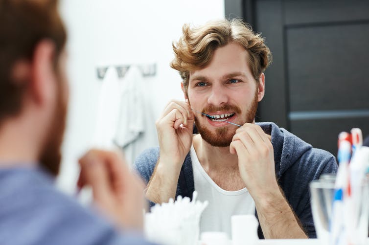 A man flosses his teeth in the bathroom mirror.