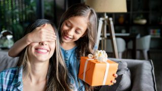 Cute little girl surprising her mom with a gift box for mother's day while covering her eyes