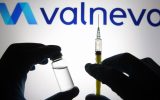 In this photo illustration, silhouette of hands in medical gloves hold a medical syringe and a vial in front of Valneva logo.