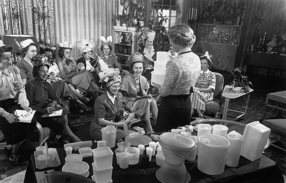 A woman holds three Tupperware containers while standing in front of a group of women seated in a living room during a Tupperware party. Some of the women wear hats made from the plastic containers. A table in the foreground displays a range of the company's products.