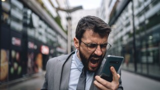 Young businessman walking outdoors in formalwear in downtown district, shouting at a mobile phone while recording a voice mail.